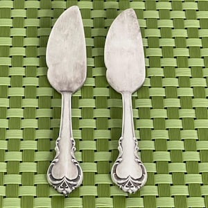 May include: Two silver cake servers with a curved blade and decorative handles. The handles feature a floral pattern at the base. The servers are displayed on a green woven placemat.