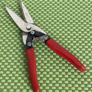 May include: Metal aviation snips with serrated silver blades and bright red rubberized handles. The tool features a central pivot bolt marked USA and WISS, with a black metal frame connecting the blades to the handles and visible screws.