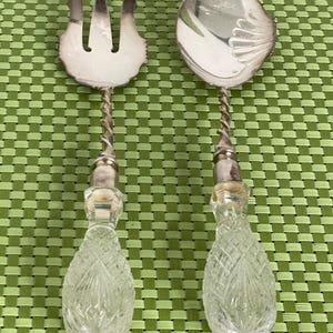 May include: A silver serving fork and spoon set. The fork has three tines, and the spoon has a shell-shaped bowl. Both have twisted silver handles and clear, cut-glass teardrop-shaped handles.