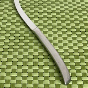 May include: A silver butter knife with a curved blade and a handle that tapers to a point. The knife is on a green woven placemat.