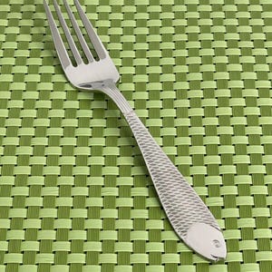 May include: A silver fork with a fish design on the handle. The fork is on a green placemat.