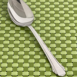 May include: A polished silver spoon with a rounded bowl and a long handle. The spoon reflects light and has a decorative end to the handle. It is set against a green woven placemat. The spoon is suitable for serving food.