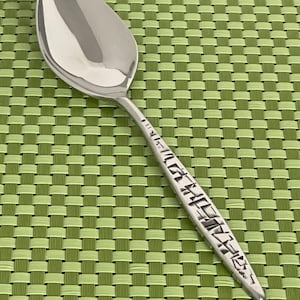 May include: A shiny, silver spoon with a rounded bowl and a long handle. The handle is decorated with a pattern of black text. The spoon is set against a green woven placemat.