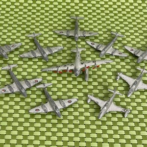 May include: A collection of small, silver-colored toy airplanes with wings and tails, arranged on a green checkered surface. The planes have detailed features, including propellers and markings.
