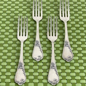 May include: Four silver forks with ornate handle detailing, arranged on a green woven placemat. The forks have a classic design with tines and decorative flourishes near the base of the handle. The overall aesthetic is vintage.