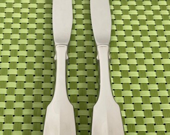 May include: Two silver butter knives with ornate handles. The knives have a polished finish and are laid out on a green woven placemat.