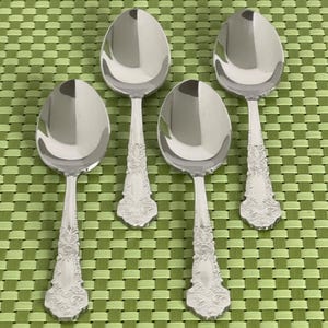 May include: Four silver spoons with ornate, detailed handles and oval bowls are arranged on a green woven placemat. The spoons are reflective, showing the surrounding environment. The spoons are likely for serving.