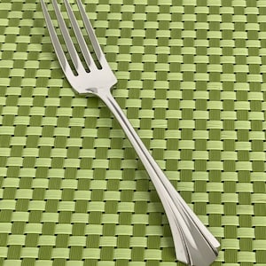 May include: A stainless steel fork with four tines, angled on a green and light green woven placemat. The fork has a long, slender handle with a decorative, geometric design at the base.