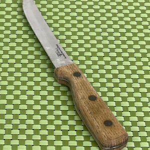 May include: A stainless steel paring knife with a wooden handle. The knife has a curved blade and a light brown handle with three black rivets. The text "Designed & Marketed by Chicago Cutlery" is visible on the blade.