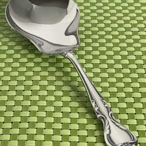 May include: A polished silver-tone serving spoon with a wide, rounded bowl and a decorative handle featuring embossed scrollwork details. The metal surface is reflective, showing a smooth finish throughout the entire length of the utensil.