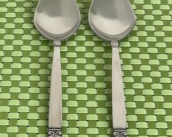 May include: Two silver serving spoons with oval bowls and decorative handles. The spoons are placed side by side on a green and white checkered surface. The handles have a detailed design at the base.