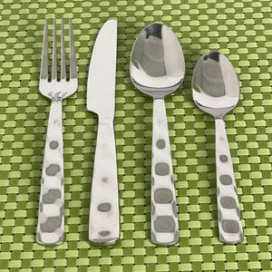 May include: A stainless steel flatware set featuring a fork, knife, and two spoons. The handles have a pattern of gray and white spots. The utensils are arranged on a green woven placemat.