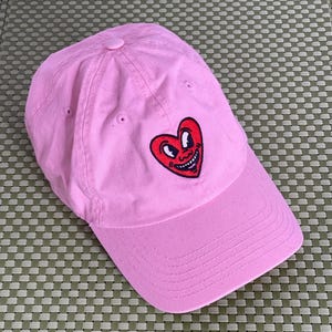 May include: A light pink baseball cap featuring an embroidered red heart graphic on the front panel. The heart design includes a cartoon-style face with large white eyes and a wide, smiling mouth with visible teeth, centered on the crown.