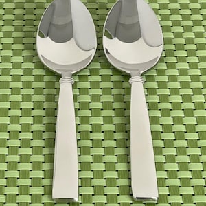 May include: Two polished stainless steel spoons with oval bowls and tapered handles. The spoons are arranged vertically against a green woven background. The spoons reflect the light, highlighting their smooth, shiny surfaces. Ideal for serving soups, stews, or other dishes.