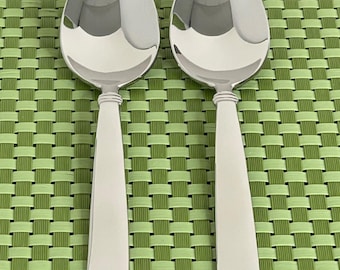 May include: Two polished stainless steel spoons with oval bowls and tapered handles. The spoons are arranged vertically against a green woven background. The spoons reflect the light, highlighting their smooth, shiny surfaces. Ideal for serving soups, stews, or other dishes.