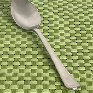 May include: A silver spoon with a rounded bowl and a long, slender handle. The spoon is resting on a green woven placemat.