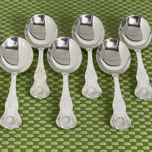 May include: Seven silver soup spoons with ornate handles are arranged on a green patterned surface. The spoons have a reflective, polished finish, and the handles feature a detailed, decorative design. The spoons are a set, likely for formal dining.