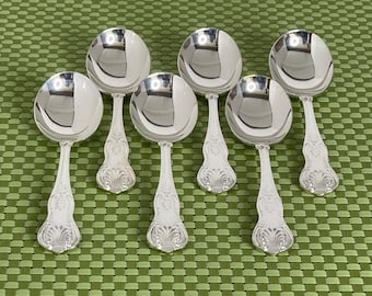 May include: Seven silver soup spoons with ornate handles are arranged on a green patterned surface. The spoons have a reflective, polished finish, and the handles feature a detailed, decorative design. The spoons are a set, likely for formal dining.