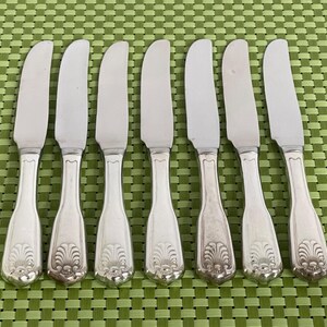 May include: Set of seven silver butter knives with curved blades and ornate handles. The handles feature a detailed, symmetrical floral design. The knives are arranged on a green checkered surface, showcasing their elegant design.