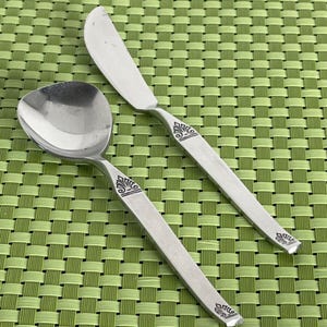 May include: Two silver utensils with ornate designs on a green woven placemat. The utensils are a butter knife and a spoon.