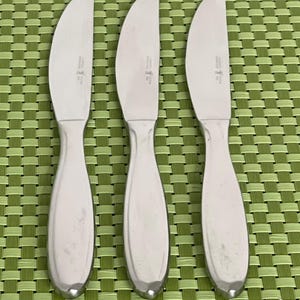 May include: Three stainless steel butter knives with rounded blades and teardrop-shaped handles. The knives are arranged on a green woven placemat. Each knife has a small emblem near the blade's base.