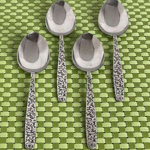 May include: Four silver spoons with an ornate floral design on the handle. The spoons are arranged in a row on a green and white woven placemat.