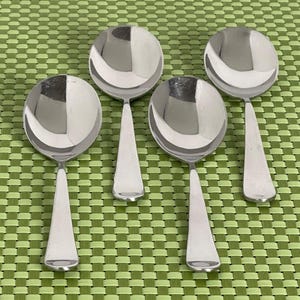 May include: Four silver soup spoons with a shiny finish. The spoons are arranged in a row on a green and white woven placemat.