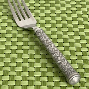 May include: A silver fork with a woven pattern on the handle. The fork is lying on a green woven placemat.