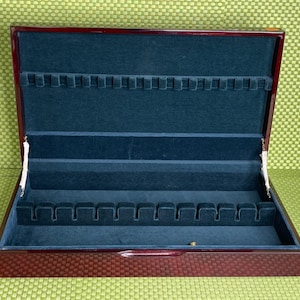May include: A rectangular wooden display case with a polished dark cherry finish and a hinged lid. The interior is lined with dark blue velvet and features two rows of slotted dividers designed to hold items securely in place.