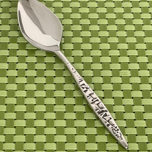 May include: A stainless steel spoon with a polished, reflective bowl and a textured handle. The handle features a repeating geometric pattern. The spoon is set against a green woven placemat.