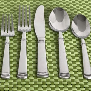 May include: A five-piece stainless steel flatware set consisting of a salad fork, a dinner fork, a dinner knife, a dinner spoon, and a teaspoon. Each piece features a smooth, polished finish with a decorative beaded band detail at the base.