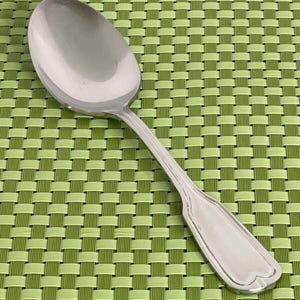 May include: A stainless steel spoon with a rounded bowl and a long, slender handle. The handle has a decorative, slightly curved end. The spoon is set against a green and light green woven placemat.