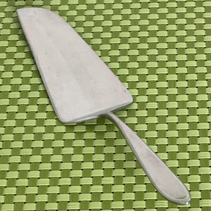 May include: A stainless steel cake server with a triangular blade and a rounded handle. The blade has a serrated edge for cutting and serving. The server is shown on a green woven placemat.