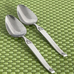 May include: Two silver spoons with ornate designs on the handles. The spoons are on a green woven placemat.