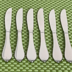 May include: Six stainless steel butter knives with curved blades and decorative handles are arranged on a green checkered placemat. The knives have a polished finish, reflecting the light.