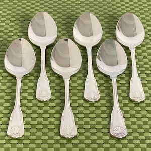 May include: Six silver spoons with oval bowls and decorative handles are arranged on a green woven surface. The spoons have a shiny, reflective surface, suggesting they are made of polished metal. The handles feature a detailed, shell-like design.