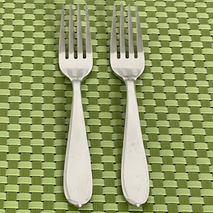 May include: Two stainless steel forks with four tines each, resting on a green woven placemat. The forks have a simple, elegant design with a smooth, rounded handle.
