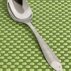 May include: A silver spoon with a fish design on the handle. The spoon is on a green placemat.