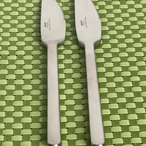 May include: Two silver butter knives with the word "IKEA" printed on the blade. The knives are on a green woven placemat.