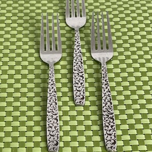 May include: Three silver forks with a decorative floral pattern on the handle. The forks are arranged in a row on a green placemat.