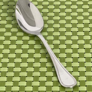 May include: A shiny, silver spoon with a rounded bowl and a detailed handle. The spoon is set against a green and light green woven background, creating a contrast in textures and colors. The spoon's design includes a beaded border.