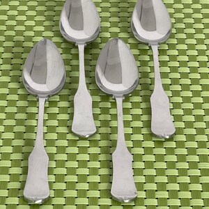 May include: Four silver spoons with oval bowls and elongated handles are arranged on a green woven placemat. The spoons have a reflective, polished surface, and the handles have a distinctive shape.