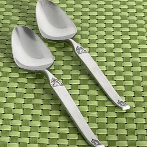 May include: Two silver spoons with ornate designs on a green woven placemat.