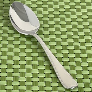May include: A shiny, silver spoon with a rounded bowl and a decorative handle. The handle features a detailed, textured design. The spoon is set against a green woven placemat.
