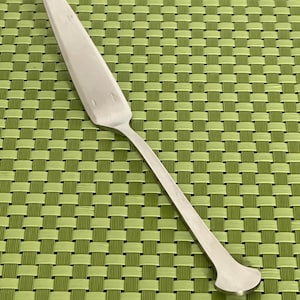 May include: A silver pie server with a long, thin handle and a wide, triangular blade. The server is made of a shiny metal and has a rounded end on the handle. The server is designed for serving pies and cakes.