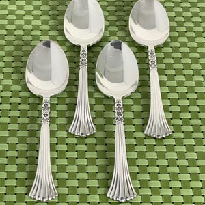 May include: Four silver-toned metal spoons arranged in a grid. Each spoon features a polished oval bowl and a handle decorated with a band of small floral motifs near the neck, transitioning into a flared, vertically fluted base at the end.