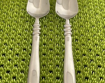 May include: Two silver spoons with rounded bowls and ornate handles. The spoons are on a green textured surface.