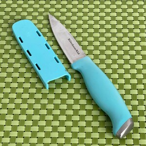 May include: A teal KitchenAid paring knife with a silver blade and handle, and a matching teal protective sheath. The knife is on a green and yellow woven surface.