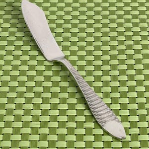 May include: A silver butter knife with a fish design on the handle. The knife is on a green woven placemat.