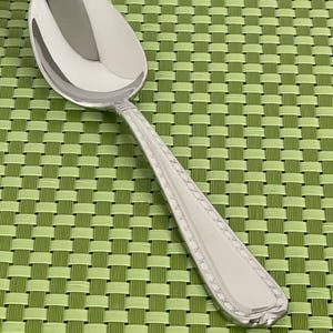 May include: A silver spoon with a decorative pattern on the handle. The spoon is lying on a green woven placemat.